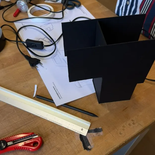 Black cardboard lamp prototype on a workbench with sketches, tools and wiring.