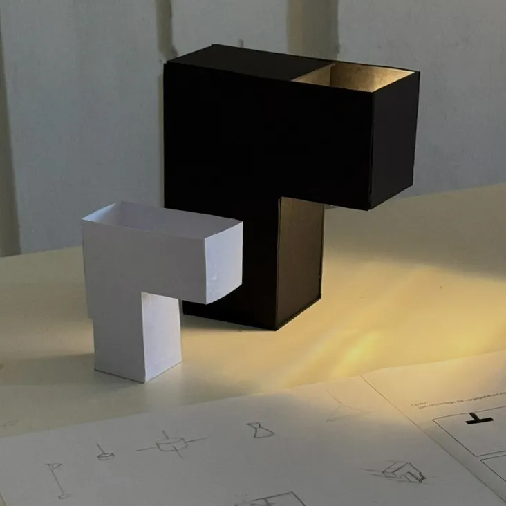 Black cardboard and white paper lamp prototypes alongside sketches, with light cast onto the table.